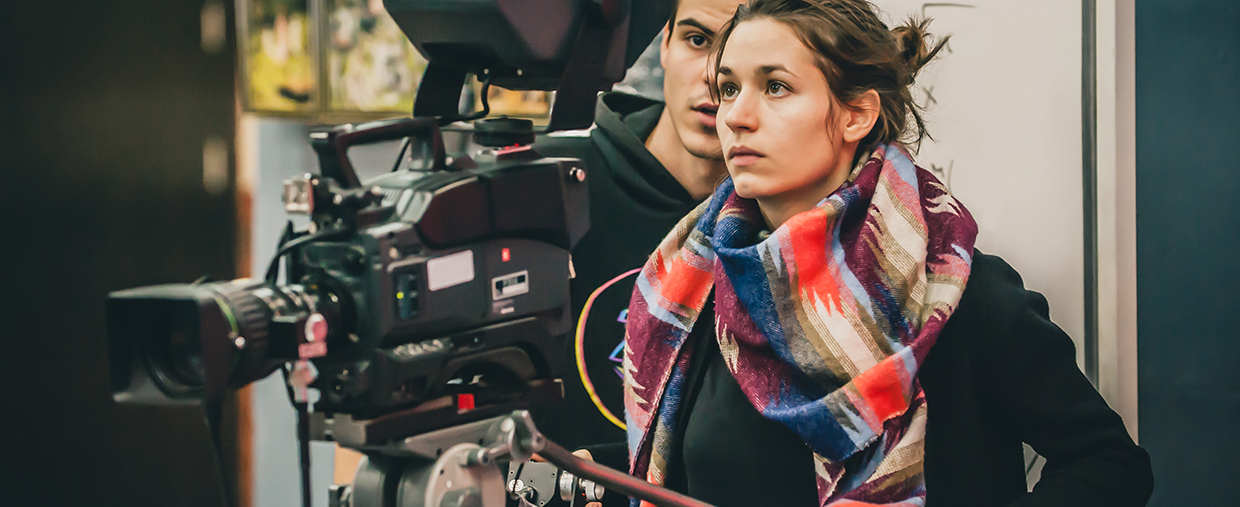 Female student is holding a large camera, filming something and staring intently at the camera's viewfinder. 