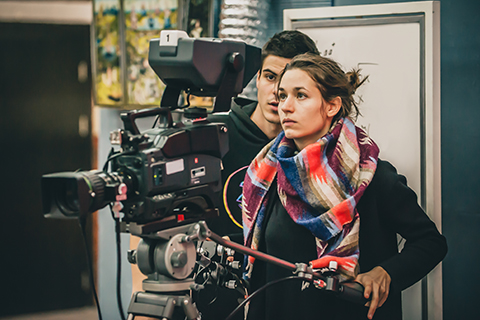 Female student is holding a large camera, filming something and staring intently at the camera's viewfinder. 